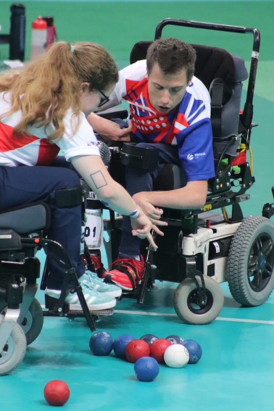 Claire Taggart and teammate David Smith, both in wheelchairs and wearing Boccia UK team kits, closely examine the position of boccia balls on the court during the 2022 World Championships team semi-final.