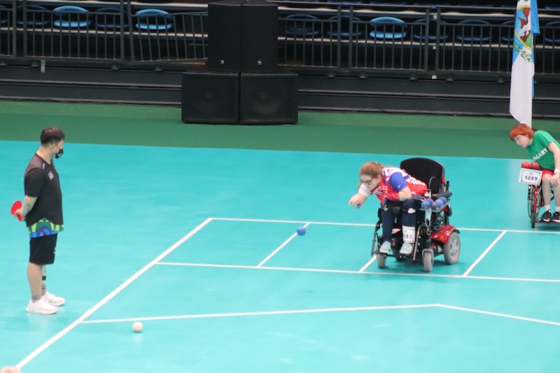 Claire Taggart, representing Great Britain, throws a blue boccia ball from her wheelchair during the 2022 World Championships semi-final, as opponents and officials observe from the court.