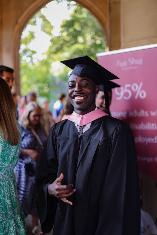 A young man in a black graduation gown and cap with a pink hood smiles confidently while making a hand gesture. He stands in front of a maroon Fair Shot banner that partially reads '95% of young adults with disabilities…'. Behind him, a crowd of people, some in summer clothing, are gathered under a stone archway with trees visible in the sunlit background.