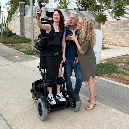 A young woman using a powered standing wheelchair takes a selfie outdoors on a paved path. She is upright in the chair and smiling, holding a smartphone on an extended grip. Two adults—a man and a woman—stand close beside her, smiling and leaning in for the photo. Trees, greenery, and a stone wall line the background on a warm, sunny day.