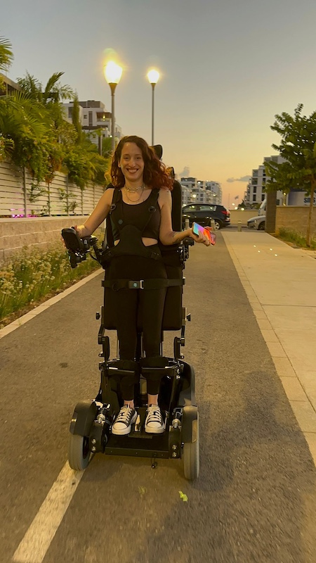 A young woman is outdoors at dusk, smiling as she uses a powered standing wheelchair on a quiet street. She is upright in the chair, wearing black clothing and sneakers, and holding colorful objects in one hand. Streetlights glow behind her, and modern buildings, trees, and a stone wall line the path as the sky transitions into evening.