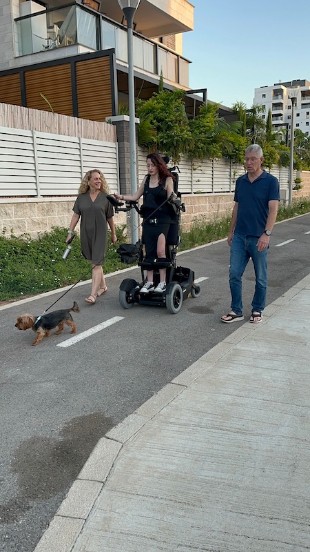 A young woman uses a powered standing wheelchair while moving along a paved path between residential buildings. She is upright in the chair and accompanied by two adults walking on either side of her—one holding a small dog on a leash. All three appear relaxed and engaged with one another as they move down the path, with greenery and modern buildings lining the walkway.