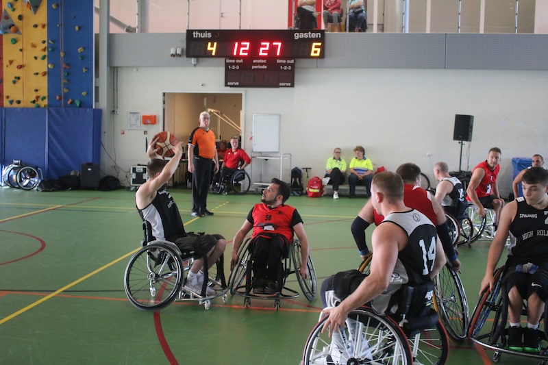 A group of men playing wheelchair basketball on a court where one is about to take a shot into the hoop. They are in two teams, one wearing red and the other wearing black and white sports kits.