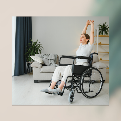 A calm, bright living room scene showing a person seated in a wheelchair, stretching their arms overhead with fingers interlaced. They are wearing light-coloured clothing and appear relaxed and focused. Behind them is a sofa with cushions and a folded blanket, a shelving unit with plants, and soft natural light coming from the left. The overall mood is peaceful and supportive, emphasizing gentle movement and comfort in a home environment.