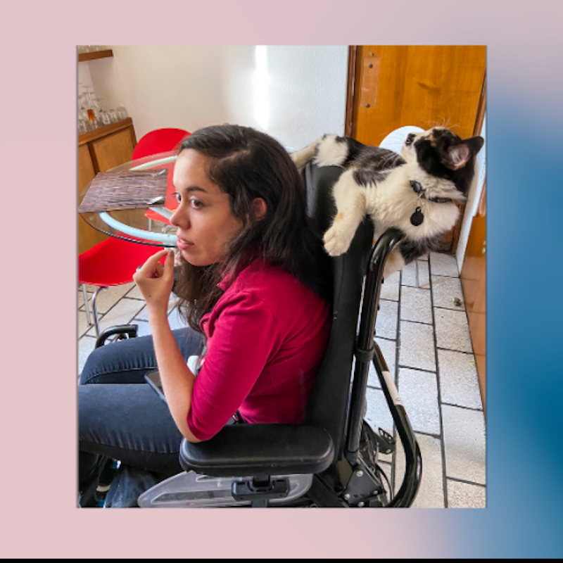 A candid indoor photo of a person seated in a wheelchair at a kitchen table, resting their chin on their hand and looking thoughtfully to the side. A black-and-white cat is sprawled comfortably across the back of the wheelchair, paws dangling and head tilted upward. In the background are red dining chairs, a wooden door, and tiled flooring. The scene feels warm and humorous, highlighting the cat’s relaxed pose and close companionship.