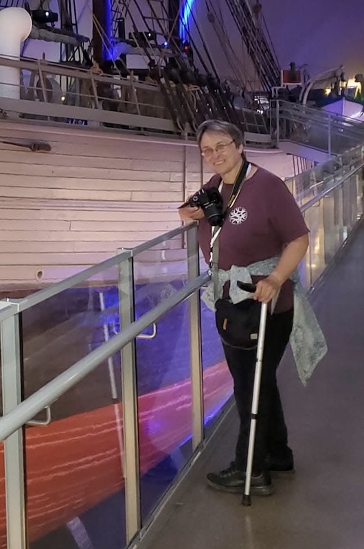 Karen smiling indoors beside a large historic ship, holding a camera and using a walking stick, standing along a glass-railed walkway with blue and purple lighting.