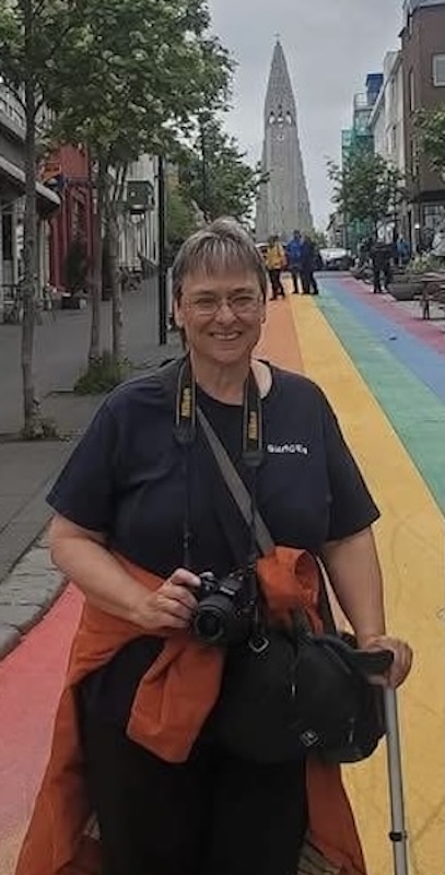 Smiling Karen holding a camera and a walking stick, standing on a rainbow-painted street with buildings and a tall church tower in the background.