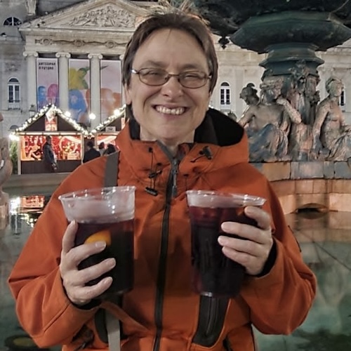 Smiling Karen wearing an orange jacket and glasses, holding two clear cups of dark drinks, standing outdoors at night in front of a lit historic building and fountain.