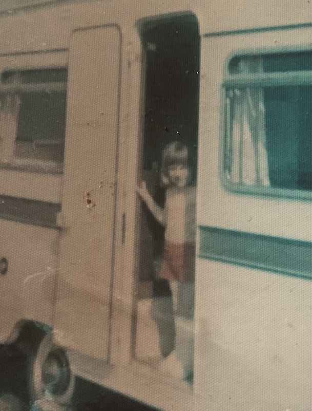 Grainy vintage photograph of Karen as a young child stepping out of a caravan doorway, wearing a light top and shorts, with the caravan exterior visible behind them.