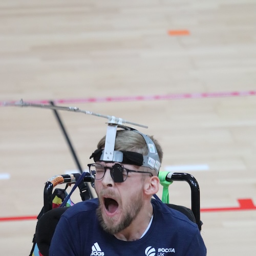 A male athlete - Patrick Wilson - in a sports wheelchair yawns or shouts during an indoor boccia match, wearing a head-mounted pointing device and dark glasses on a wooden court with court markings visible in the background.