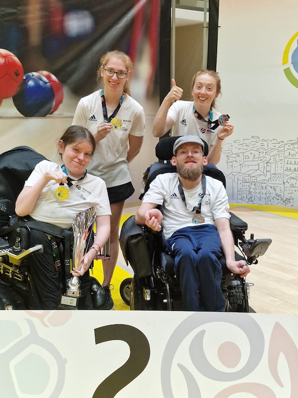 Patrick Wilson and Sally Kidson sit in powered wheelchairs holding medals, joined by ramp operators Chloe Lewis and Meredith Greenwood standing behind them, all smiling and celebrating. The group celebrate silver at the Coimbra World Cup, with medals visible, one person giving a thumbs-up, and a sports venue backdrop featuring boccia imagery.