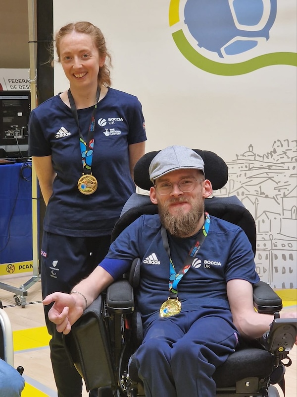 Meredith Greenwood stands smiling, while Patrick Wilson sits in a powered wheelchair, also smiling, with a medal around their neck. A competition banner and sports venue details are visible in the background.”