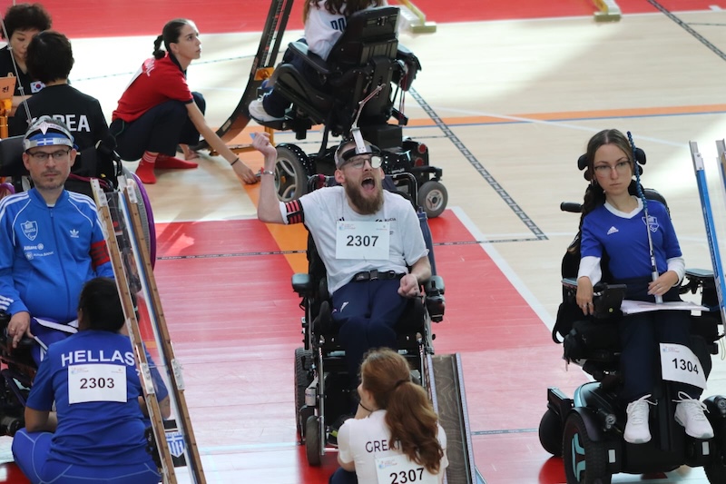 Patrick Wilson celebrates a successful shot during an indoor boccia match, raising his arm and shouting while seated in a powered wheelchair on a marked court. Other boccia players, referees, and assistants are visible nearby, with competition numbers and team uniforms shown in the background.
