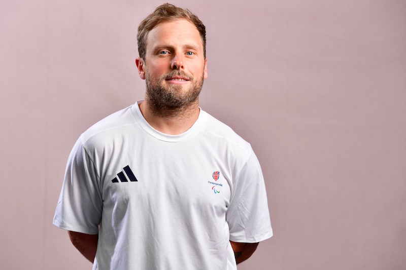 Portrait of James Barnes-Miller looking at the camera against a soft pink background. He is wearing a white short-sleeved Team ParalympicsGB T-shirt featuring the ParalympicsGB logo on the chest and an Adidas logo on the opposite side, with his hands held behind his back.