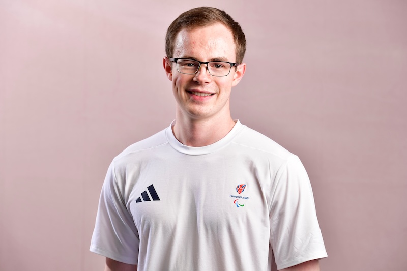 Portrait of Neil Simpson smiling at the camera against a soft pink background. He is wearing a white short-sleeved Team ParalympicsGB T-shirt with the ParalympicsGB logo on the chest and a small Adidas logo on the opposite side.