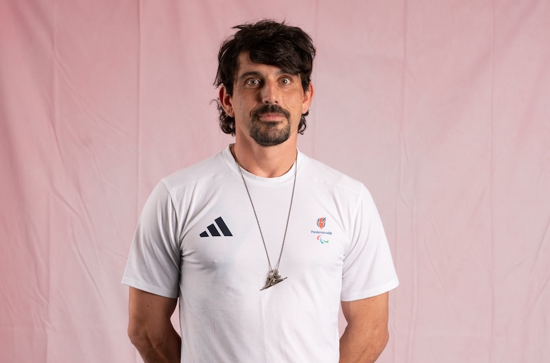 Portrait of Ollie Hill facing the camera against a soft pink background. He is wearing a white short-sleeved Team ParalympicsGB T-shirt with the ParalympicsGB logo on the chest and an Adidas logo on the opposite side. He also wears a necklace with a pendant, and has dark hair and facial hair.