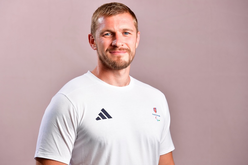 Portrait of Scott Meenagh smiling slightly at the camera against a soft pink background. He is wearing a white short-sleeved Team ParalympicsGB T-shirt featuring the ParalympicsGB logo on the chest and a small Adidas logo on the opposite side.