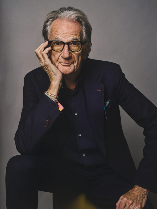 Portrait of fashion designer Sir Paul Smith seated against a plain grey background, wearing dark clothing and glasses, resting his face on one hand while looking directly at the camera.