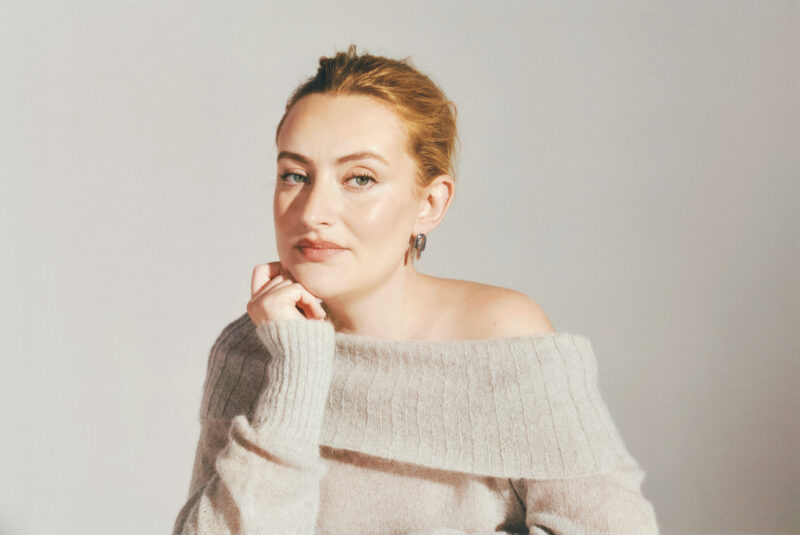 Portrait of presenter and producer Amelia Dimoldenberg wearing a soft off-the-shoulder knit jumper, resting her chin on her hand and looking towards the camera against a light neutral background.