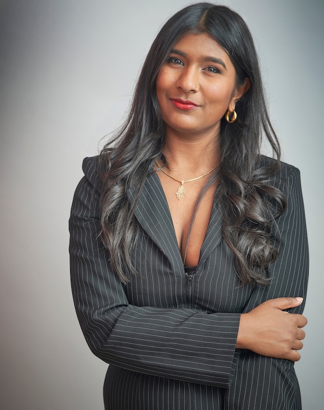 Portrait of writer and broadcaster Ash Sarkar wearing a dark pinstripe blazer, standing with folded arms and looking towards the camera against a neutral grey background.