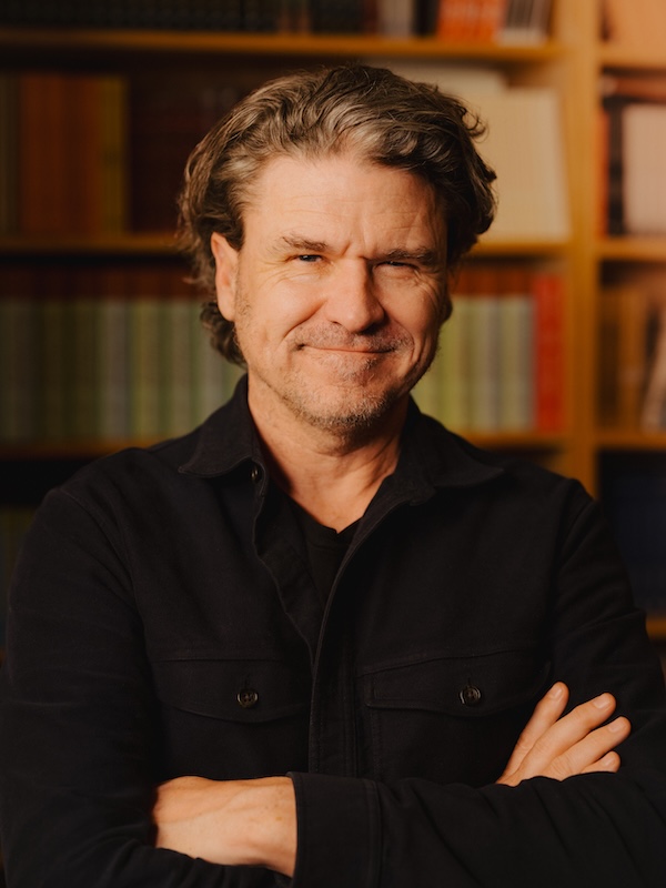 Portrait of author Dave Eggers standing with folded arms in front of bookshelves, wearing a dark jacket and smiling slightly towards the camera in warm lighting.