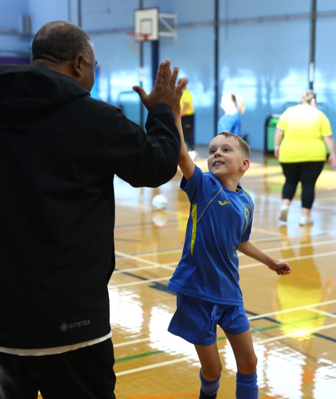 A young player in a blue football kit celebrates with a high-five from a coach during an indoor pan-disability football training session, with other participants and football activities taking place in the background.