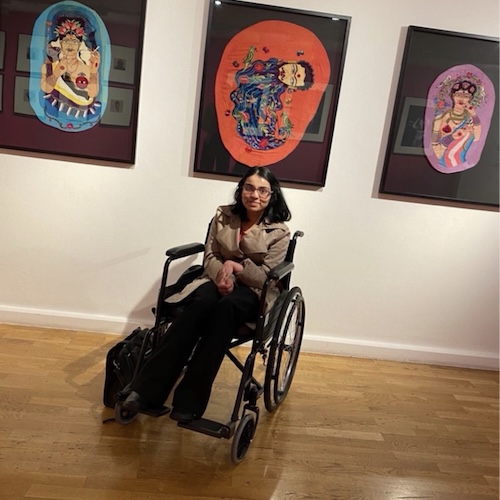Gina Patel sits in a wheelchair in an art gallery, positioned beneath three framed colourful portraits on a white wall, with a wooden floor and exhibition space surrounding her.