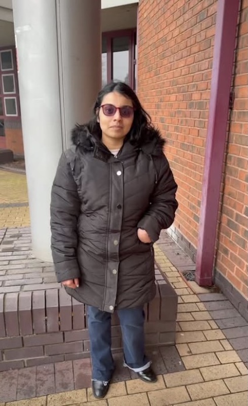 Gina Patel stands outdoors in front of a brick building, wearing a black winter coat and glasses, with one hand in her pocket.