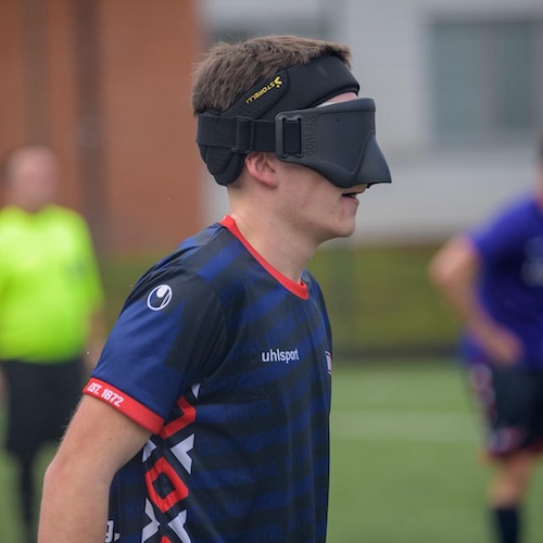 Side profile of blind footballer Olly Johnson wearing an RNC Hereford kit and protective eye shades during a match, with a referee and opposing player blurred in the background.