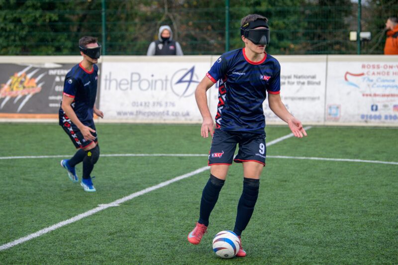 Olly Johnson controls the ball during a blind football match for RNC Hereford, wearing protective eye shades and the club's navy-blue kit, with a teammate nearby on the pitch.