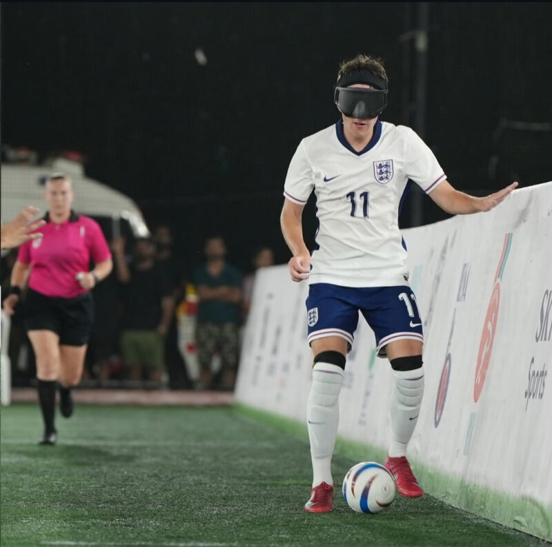 Olly Johnson represents England in blind football, dribbling the ball along the touchline while wearing the England number 11 shirt and protective eye shades during an international match.