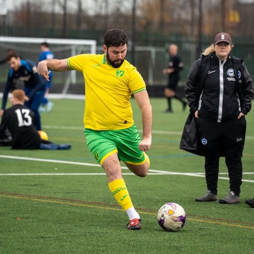 Owen Woodrow, wearing Norwich City’s yellow and green kit, controls the ball during a cerebral palsy football training session or match on an outdoor artificial pitch, with teammates and coaching staff visible in the background.